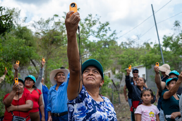 Group of people outdoors hold up small orange handheld devices toward the sky, likely monitoring weather conditions, with trees, buildings and cloudy sky in background.