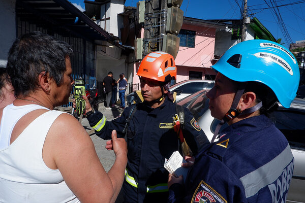 Emergency responders in helmets talk with a resident on a street lined with damaged buildings, vehicles and overhead cables.