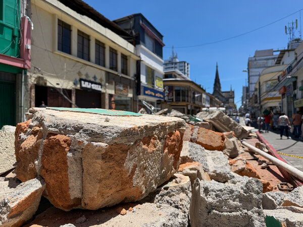 Rubble from collapsed masonry blocks part of a city street after an earthquake, with damaged buildings and pedestrians visible beyond.