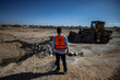 A man in an organ vest with a WFP logo on its back observes a construction tractor scooping rocks on a desert road