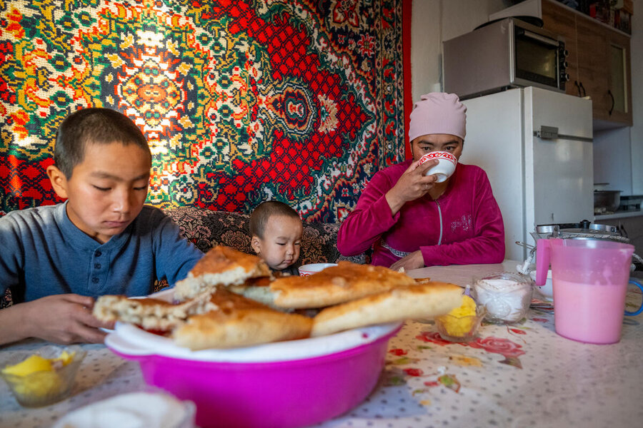 A woman and two young boys sat at a table in front of food. There is a bowl of bread in the foreground and the woman is drinking from a cup.