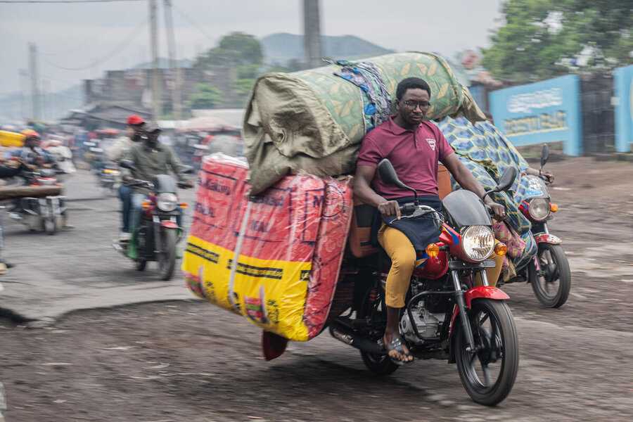 A tired looking man on a motorcycle laden with a double mattress rides by, followed by others