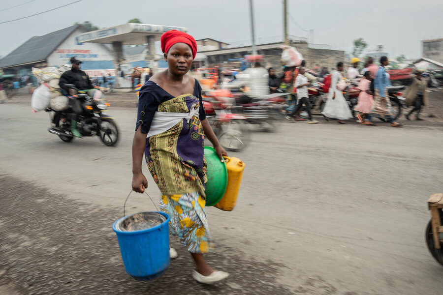 An evacuating woman in a red headscarf carries a bucket as a motorcycle laden with belongings passes by