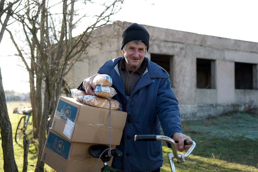 An older gentleman wearing a black woolly hat, wheels two WFP boxes and bread on his bicycle