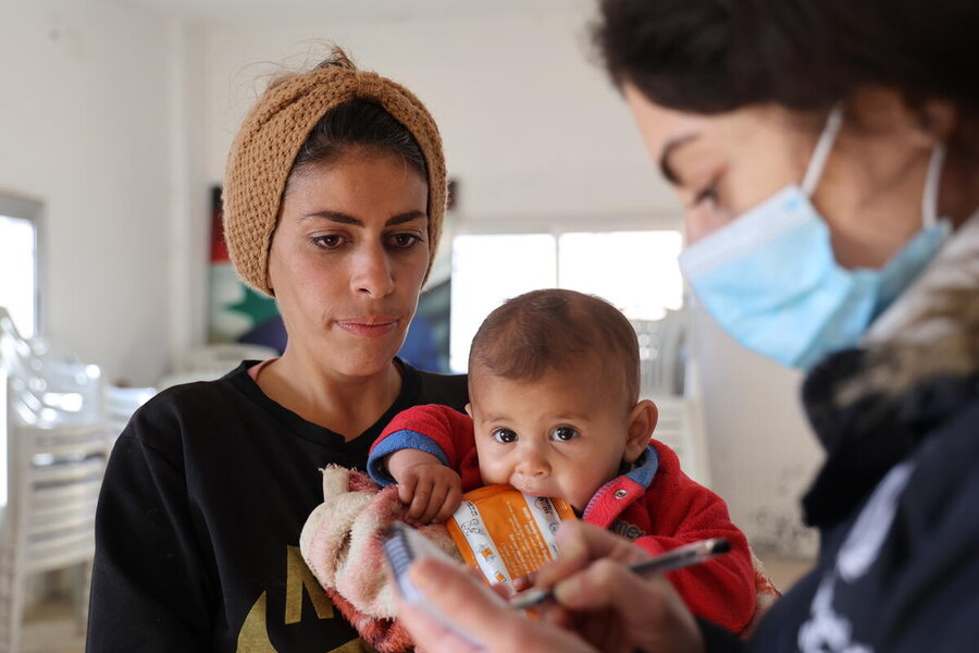 A baby looks into camera, held by her mother, as nutritionist looks on wearing a facemask