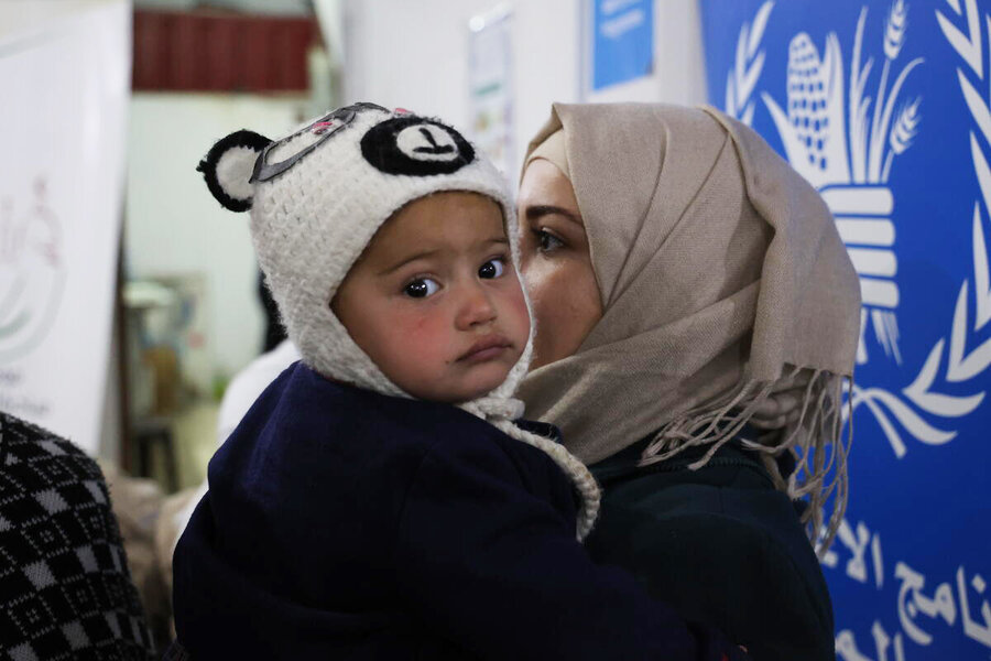 A toddler is held by their mother who is sat in front of a big blue WFP sign