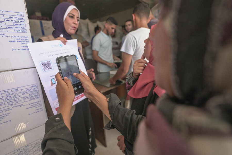 A person scans a QR code on a printed notice using a mobile phone at a crowded assistance site, while others wait near a counter indoors.
