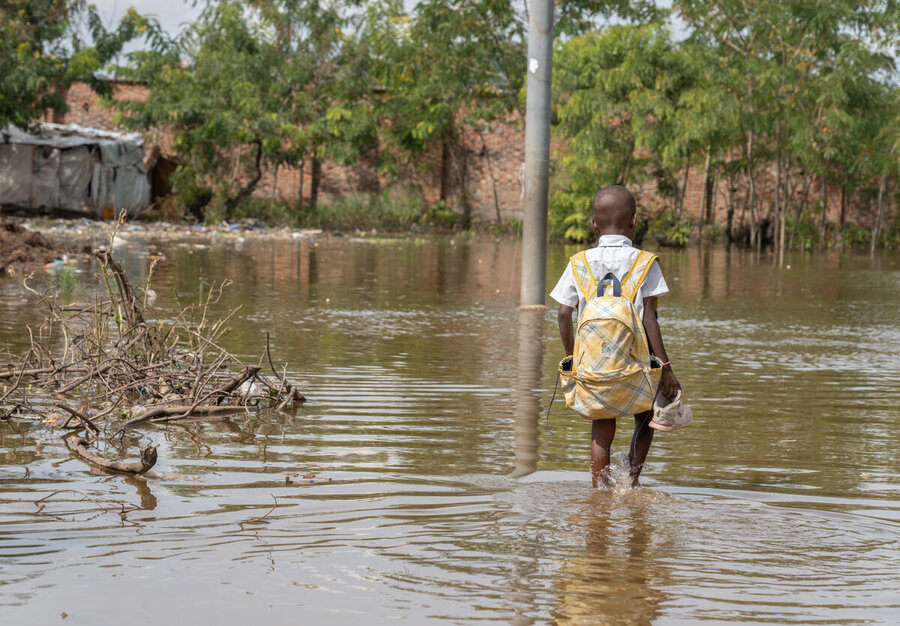 Child with backpack walks through knee‑deep floodwater, holding shoes, as homes, trees and debris line a flooded street in a damaged neighbourhood.