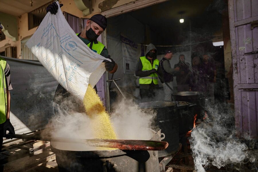 A man sporting a green vest,black cap and facemark pours grains into a large, black boiling pot. Photo: WFP/Jaber Badwan